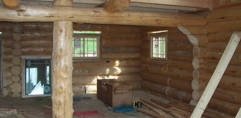 Here's a concise and descriptive alt text for the image: Interior view of a log cabin under construction, showing peeled log walls, a central support column