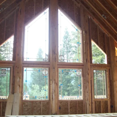 Interior view of a wooden frame house under construction, showing a large, two-story wall of windows in a triangular arrangement looking out onto a forest of pine trees.
