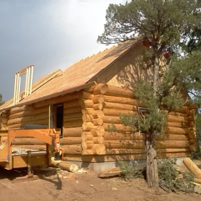 Exterior shot of a log cabin under construction, featuring a peeled log structure, an unfinished roof with OSB sheathing, a visible second-story dormer frame, and an orange trailer parked on the dirt construction site near a large pine tree.