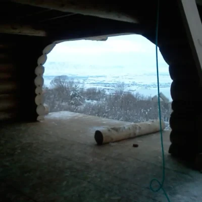 Interior shot of a dark log cabin under construction, showing a large, rounded opening cut into the log wall that offers a panoramic view of a snow-covered valley and mountains.