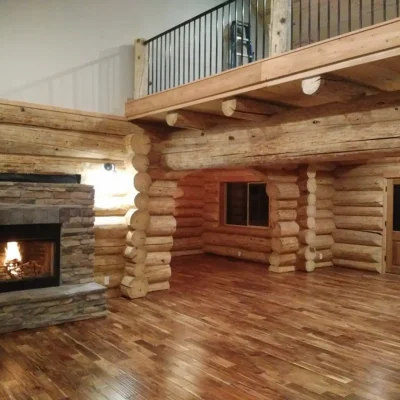 Interior of a finished log cabin great room, with a stone fireplace, wood floor, and a second-story loft featuring peeled log construction and black railings.