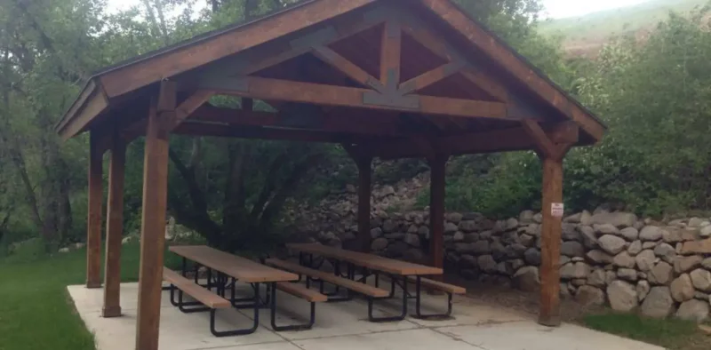 An outdoor wooden picnic pavilion with a dark-stained roof structure, covering a concrete pad with two long wooden picnic tables, set on a grassy lawn next to a low stone retaining wall and surrounded by trees.