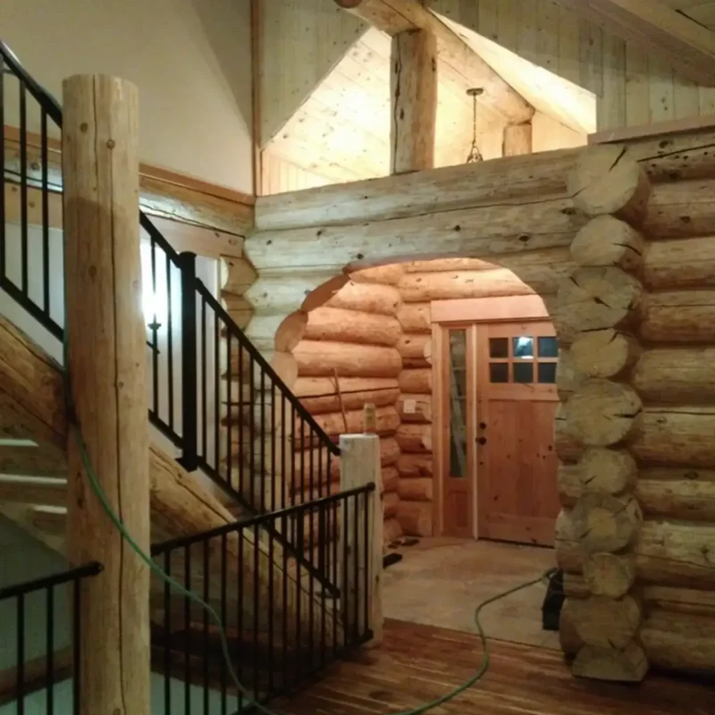 Interior view of a rustic log cabin showing a staircase with black metal railings, a large peeled log support post, an arched doorway cut into the thick log wall, and a wood plank door in the background.