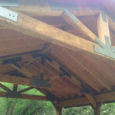 A close-up, upward view of the exposed wooden truss work and ceiling beams of an outdoor pavilion, featuring large, decorative black metal plates and bolts used to join the timber members.
