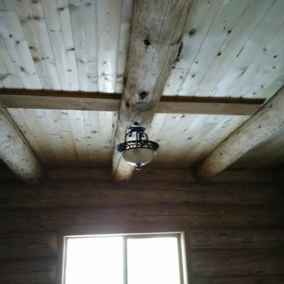 An upward interior view of a rustic ceiling featuring exposed peeled log beams, natural wood planking, and a single decorative bowl-style light fixture hanging from the center.