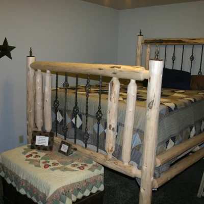 A rustic bedroom features a bed frame made of peeled logs with black metal spindles, covered with a quilted bedspread. A dark storage chest with a quilt is at the foot of the bed, and two decorative metal stars hang on the light blue wall.
