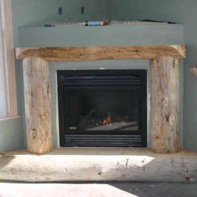 An unfinished corner fireplace in a room with light green walls, featuring a black gas insert framed by a rustic mantel and hearth made of large, peeled logs, and flanked by two large windows.