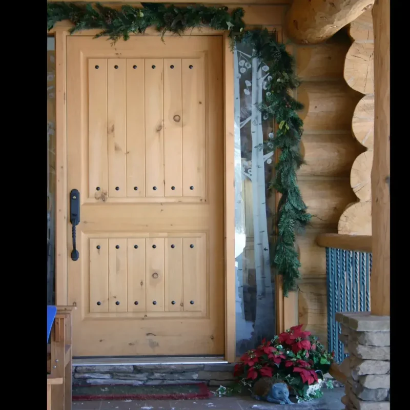 A rustic front entrance of a log cabin, featuring a light-colored wooden door with vertical planks and decorative black pegs, flanked by a narrow window and thick log wall, all decorated with evergreen garlands and a poinsettia plant.