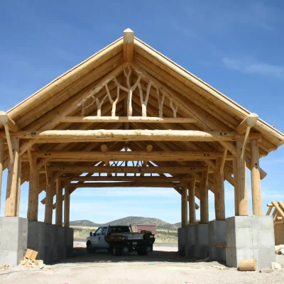 An open-air wooden log pavilion under construction, featuring a tall, peeled log frame resting on concrete foundation walls and distinctive, decorative branched log trusses visible in the gable end, with a pickup truck parked inside.