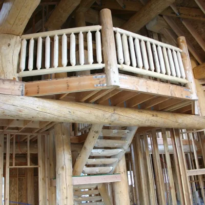Interior of a log cabin under construction, featuring an exposed wood-framed wall structure, a rustic wooden staircase, and a loft with a balcony railing made of peeled log balusters.