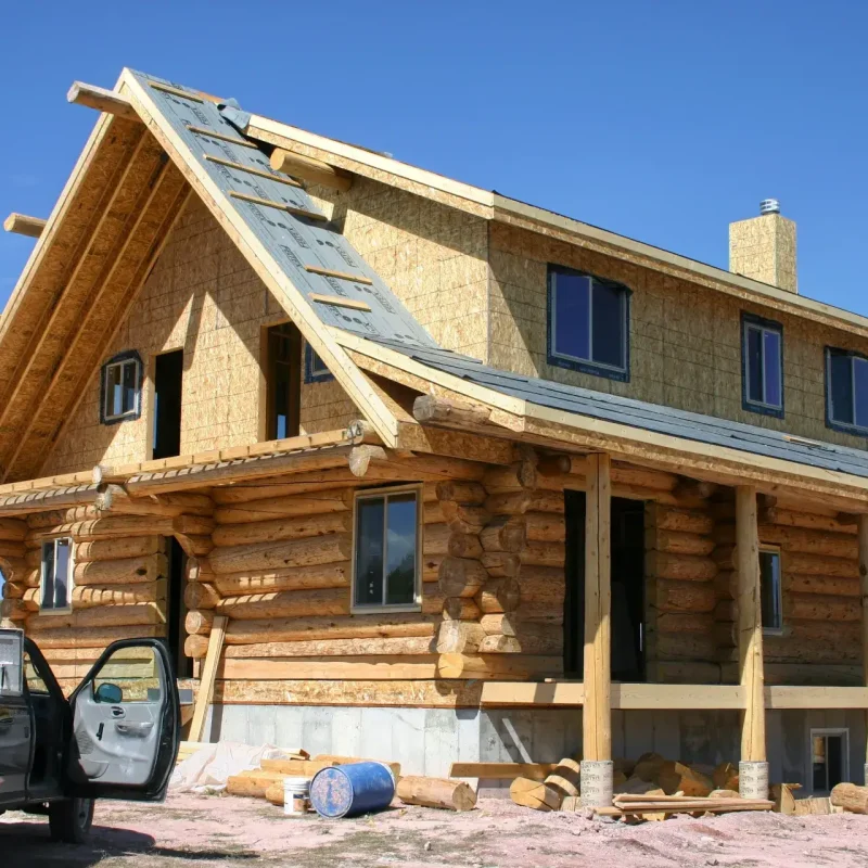 Exterior view of a large log home under construction, featuring peeled log walls on the lower level, a covered porch supported by log posts, a second story sheathed in OSB, and an unfinished gable roof, all under a bright blue sky.