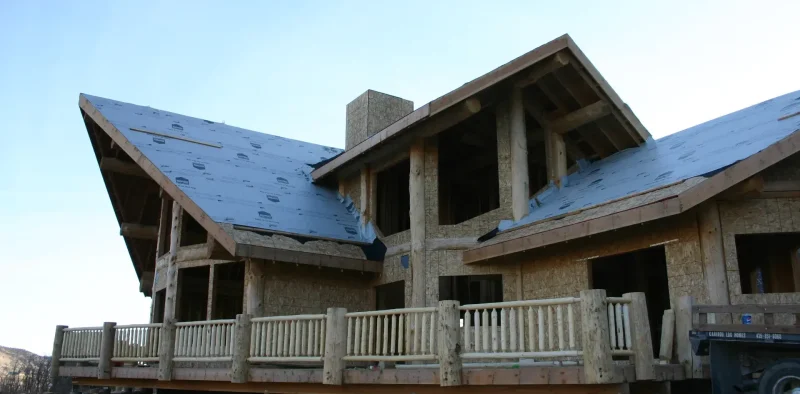 Exterior view of a large timber-frame and log home under construction, featuring multiple gables, exposed wooden framing, sheathed walls, and a deck with a rustic railing made of peeled logs, all beneath a light blue sky.