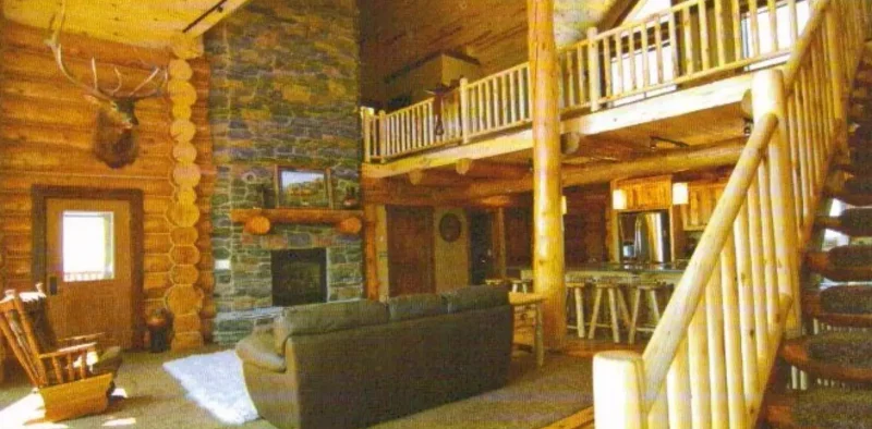 Interior of a grand log home great room, featuring a tall, stone fireplace, a staircase and second-floor loft with peeled log railings, a high vaulted ceiling, and a large mounted elk head above the main floor entrance.