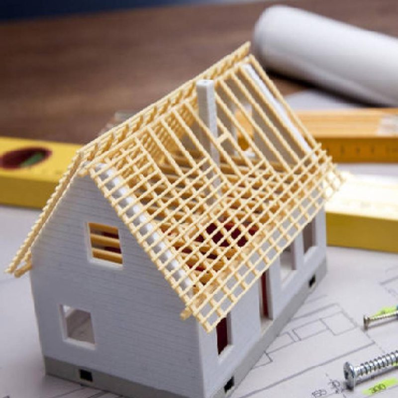 A miniature house model with a partially constructed wooden roof sits on architectural blueprints, surrounded by construction tools, screws, and a measuring level.